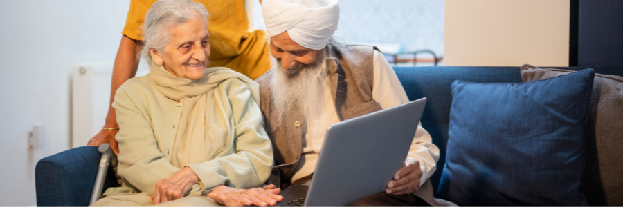 Two older people sitting on a sofa. They are smiling as the look at a open laptop that is resting on their knees.
